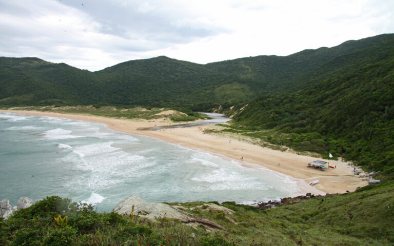 Breiter Sandstrand in Bucht mit Wasserzufluss aus den umliegenden grünen Hügeln