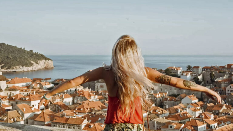 Blonde Frau mit Tattos und ausgestrecken Armen blickt auf Dubrovnik (Ansicht von hinten)