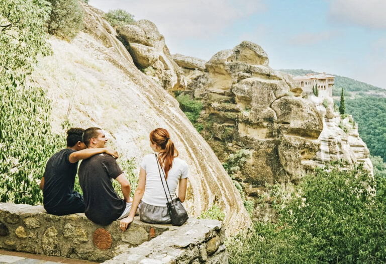 3 Reisende sitzen auf einer Mauer und blicken auf Meteora