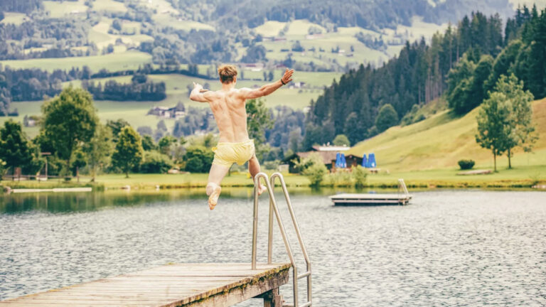 Junger Typ springt vom Steg mit ausgestrecckten Armen in den See