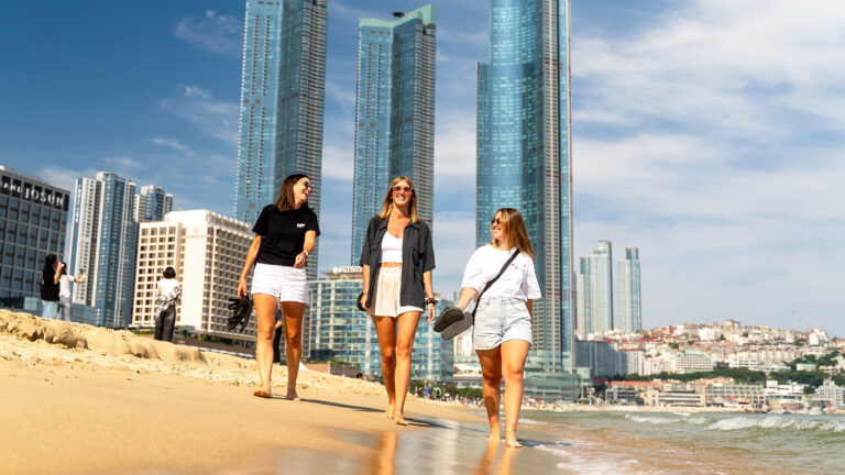 3 Mädels spazieren am Strand von Busan mit verglasten Hochhäusern im Hintergrund